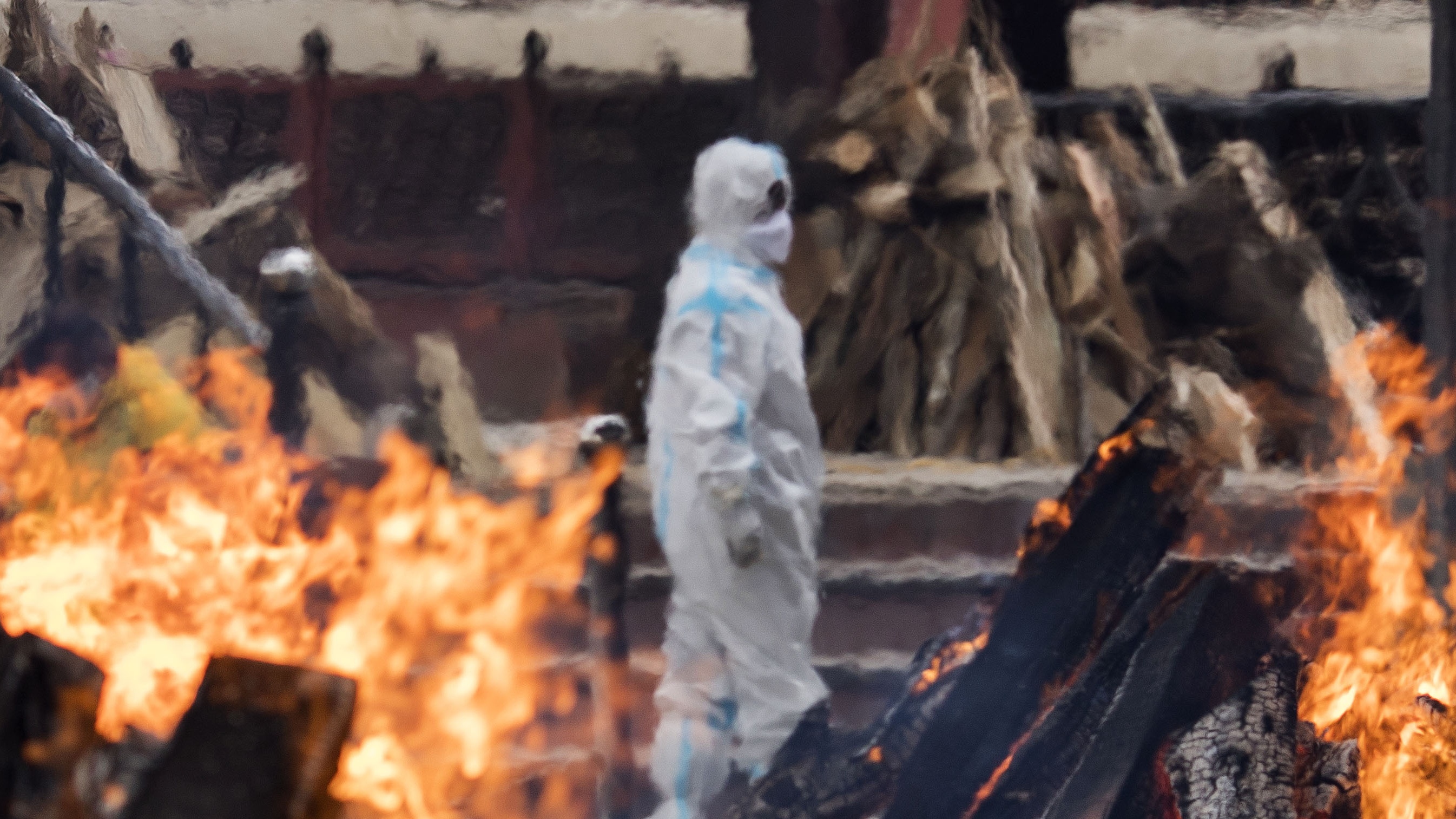 A man in PPE looks on as funeral pyres burn