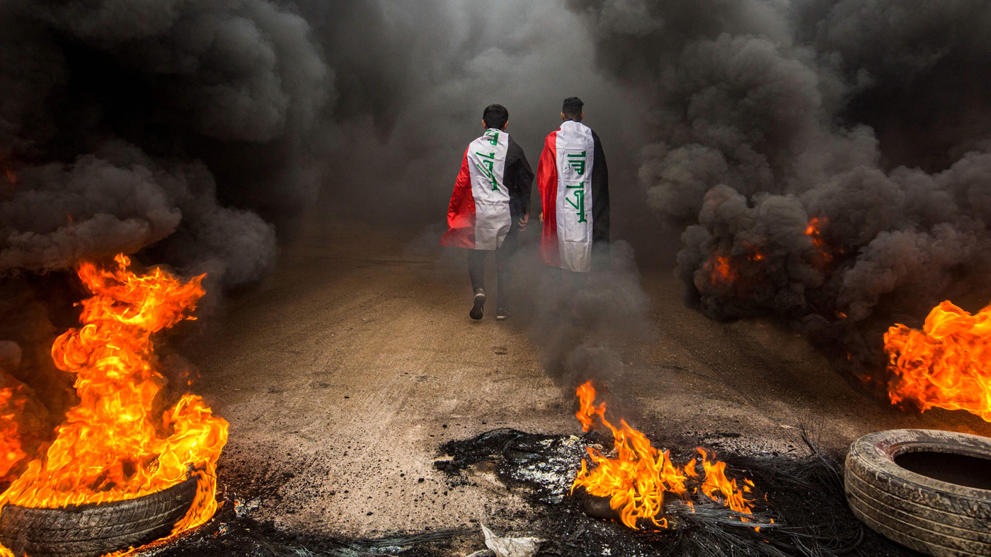 TOPSHOT - Anti-government protesters draped in Iraqi national flags walk into clouds of smoke from burning tires during a demonstration in the southern city of Basra on November 17, 2019, as 