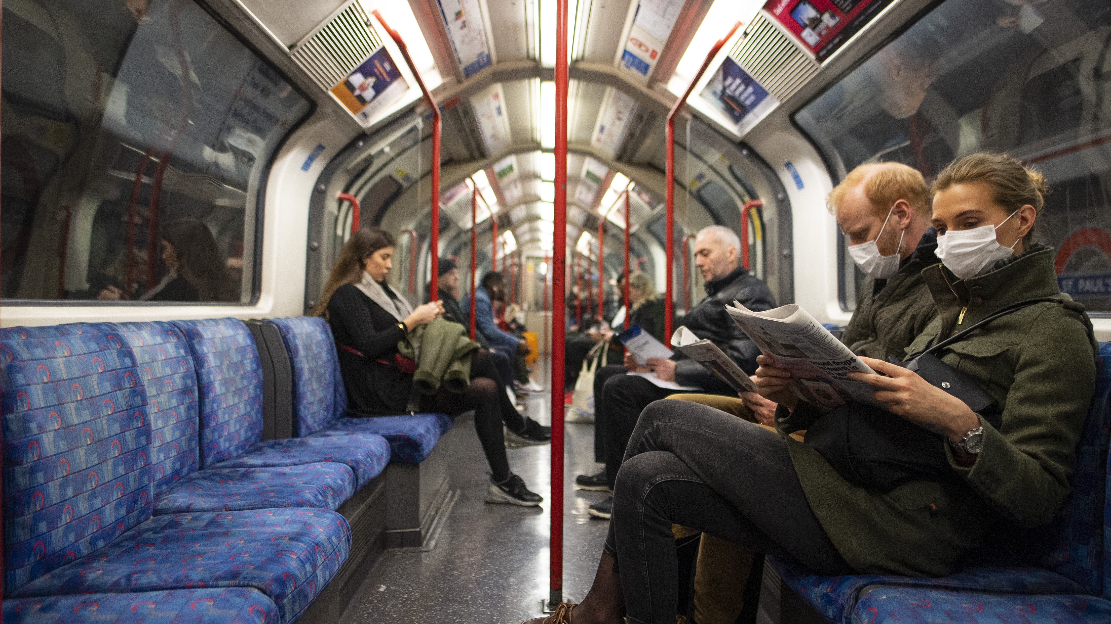 A couple sit on the Central Line Tube wearing protective face masks while reading a newspaper.