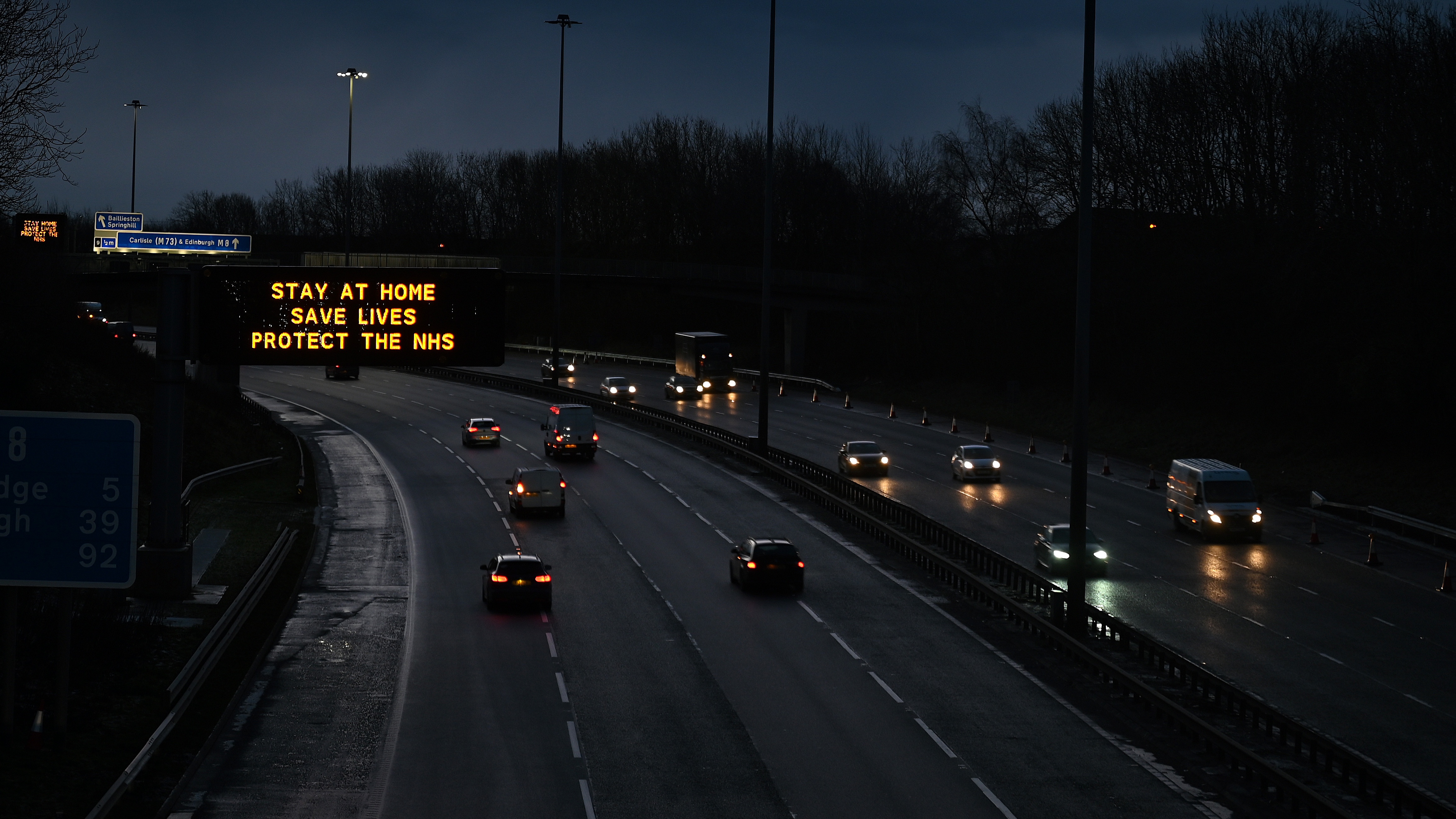 Traffic on the M8 motorway passes a road sign advising motorists to stay at home