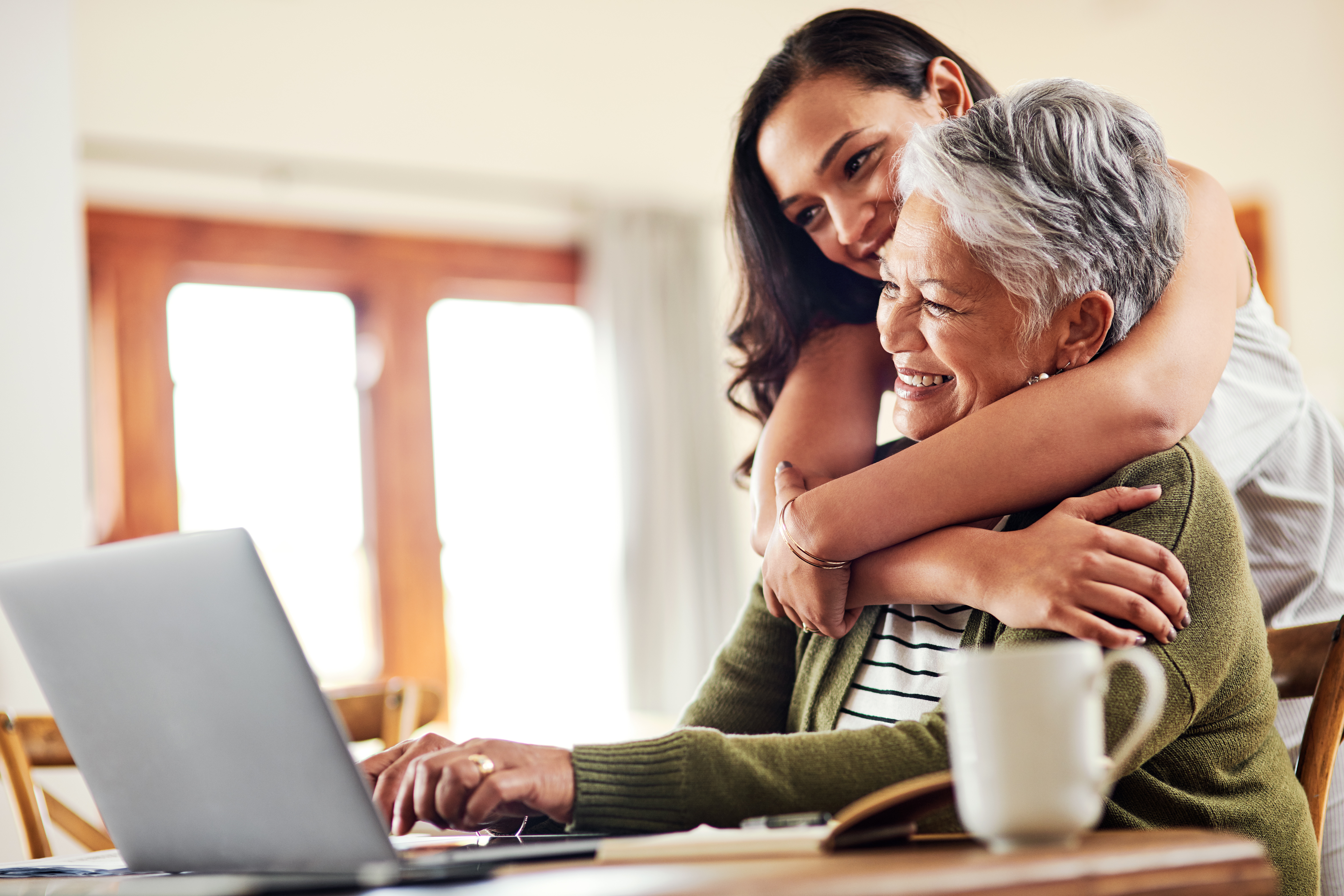 An older woman sat at a laptop with a younger woman hugging her from behind