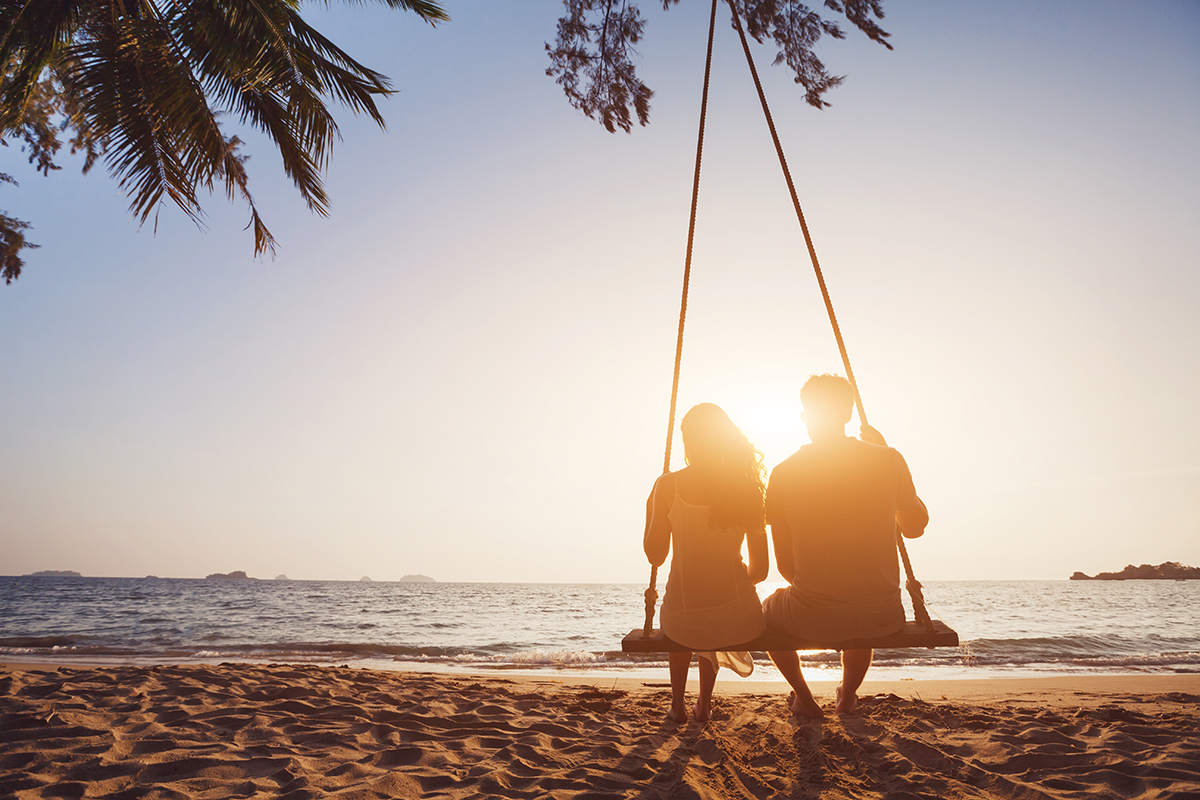 A woman and man sat on a swing on a beach. The sun is going down.